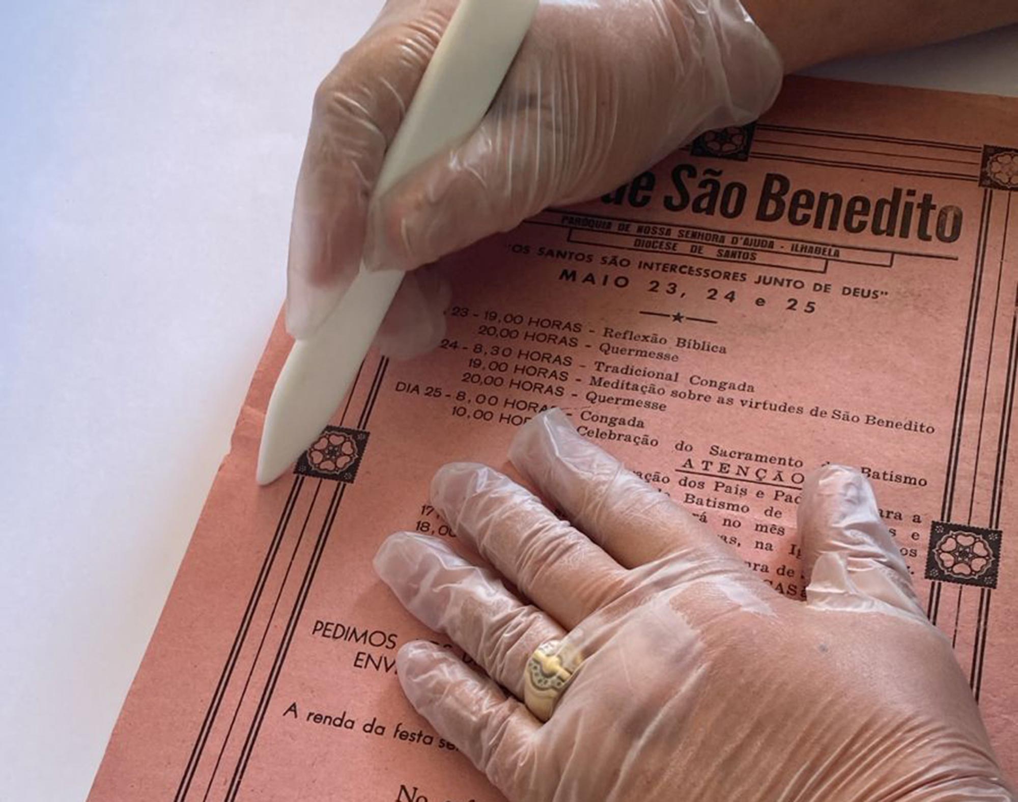 Arquivista da casa do Patrim&ocirc;nio veste luvas e manuseia um documento antigo com uma esp&eacute;cie de caneta usada para tirar vincos. A imagem &eacute; fechada e mostra detalhes das m&atilde;os e do documento.