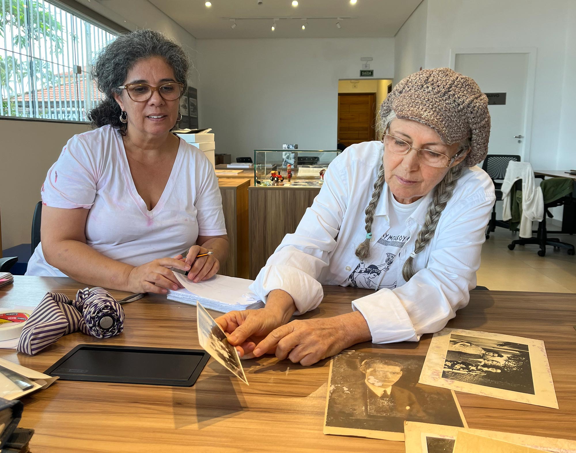 Duas mulheres est&atilde;o sentadas na frente de uma mesa na Casa do Patrim&ocirc;nio, em S&atilde;o Sebasti&atilde;o. Sobre a mesa est&atilde;o dispostas diversas fotografias em preto e branco. As duas vestem branco. A da esquerda &eacute; parda e usa &oacute;culos. A da direita usa duas tran&ccedil;as e uma boina e est&aacute; segurando uma foto.