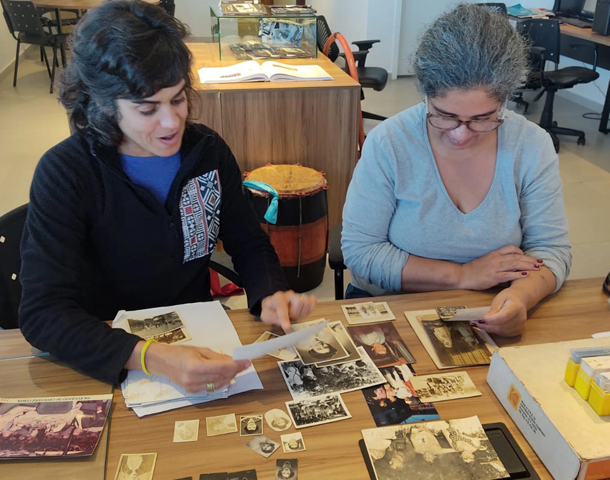 Na Casa do Patrim&ocirc;nio de S&atilde;o Sebasti&atilde;o, duas mulheres observam fotografias em cromo dispostas sobre uma mesa. A da esquerda tem cabelos curtos e veste um casaco preto. A da direita &eacute; parda, usa &oacute;culos e veste uma blusa cinza.