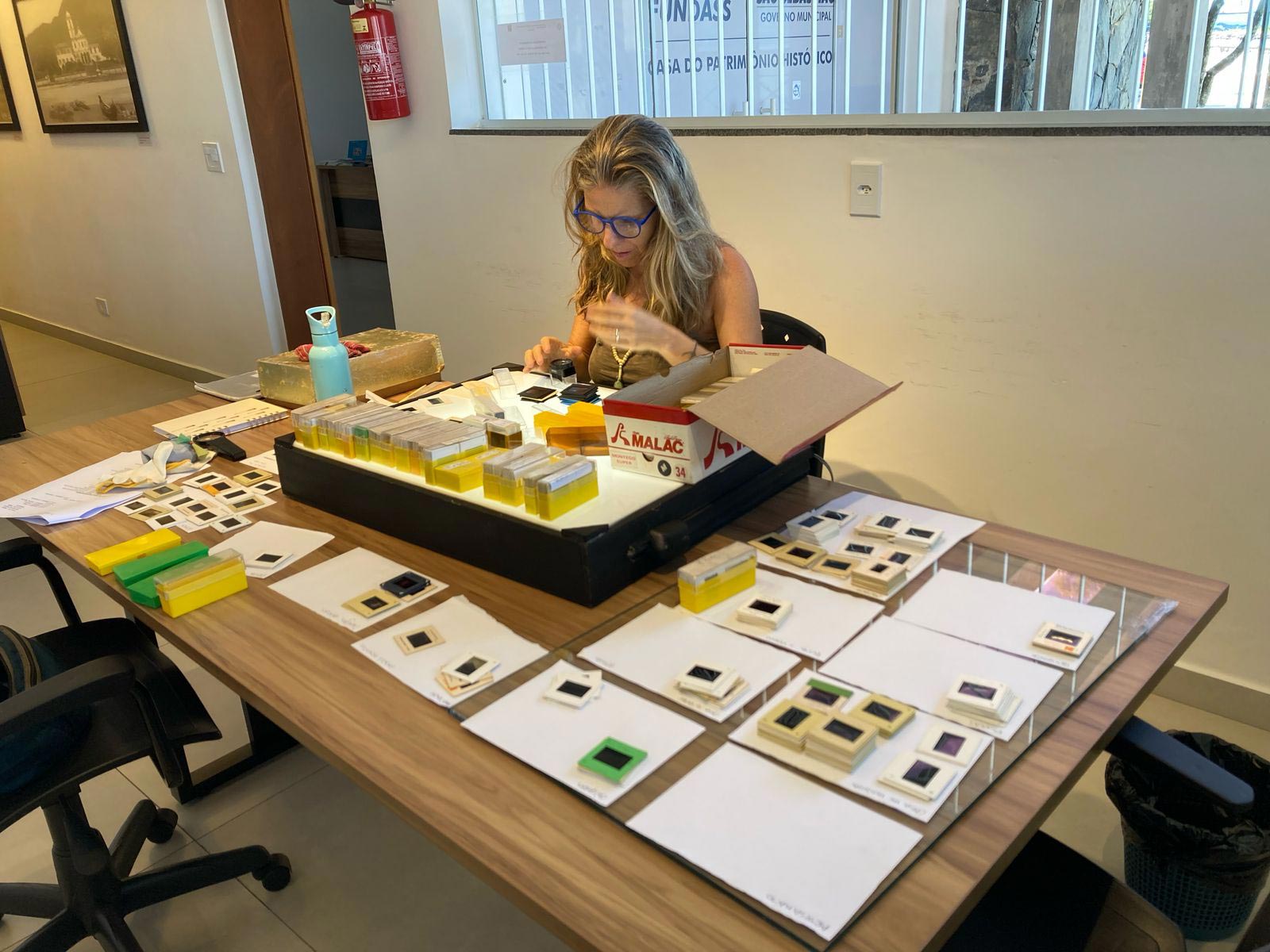 Na Casa do Patrim&ocirc;nio, uma mulher est&aacute; sentada na frente de uma mesa examinando fotografias em slide sobre uma mesa de luz. Ao redor da mesa de luz h&aacute; v&aacute;rias folhas de papel com slides dispostos de maneira organizada sobre elas.