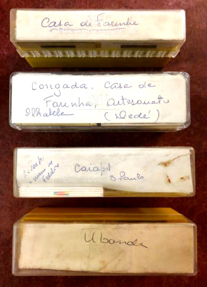 Fotografia de quatro caixinhas de guardar slides dispostas simetricamente, identificadas com escritos feitos &agrave; m&atilde;o. Na primeira, de baixo para cima, l&ecirc;-se &ldquo;Casa de Farinha&rdquo;; na segunda, Congada, Casa de Farinha, Artesanato Ilhabela, Ded&eacute;. Na terceira caixinha l&ecirc;-se &ldquo;Caiap&ocirc; S&atilde;o Paulo&rdquo; e na &uacute;ltima, &ldquo;Ubanda
