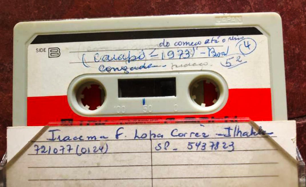 Imagem de fita K7 branca e vermelha, onde se l&ecirc; escrito &agrave; m&atilde;o: Caiap&ocirc; 1973 do come&ccedil;o at&eacute; o meio, congada peda&ccedil;o, 52. Na parte inferior h&aacute; um papel tamb&eacute;m escrito &agrave; m&atilde;o, onde se l&ecirc;: Iracema F. Lopes Corr&ecirc;a - Ilhabela, 721077, SP 54307823.