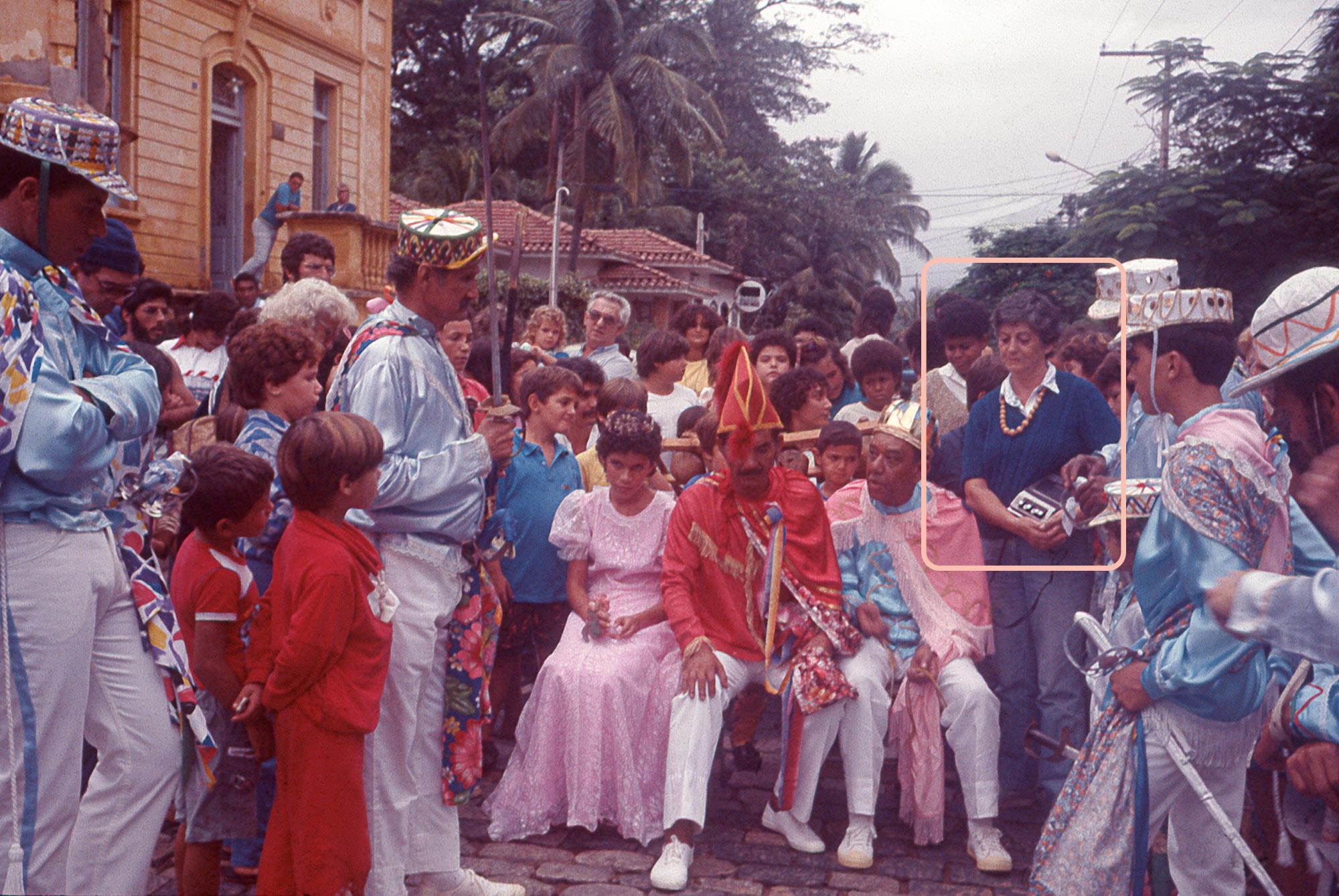 Na apresenta&ccedil;&atilde;o da Congada de S&atilde;o Benedito, um grupo de pessoas de p&eacute; est&aacute; rodeando tr&ecirc;s pessoas sentadas: a Rainha, que &eacute; uma menina com vestido rosa e uma coroa, o Embaixador, que &eacute; um homem mais velho vestindo uma farda vermelha com chap&eacute;u vermelho e cal&ccedil;as brancas, e o Rei, um homem tamb&eacute;m mais velho com uma coroa de rei, uma capa rosa e uma farda azul com cal&ccedil;a branca. Ao lado do rei, de p&eacute;, segurando um gravador, est&aacute; Iracema Fran&ccedil;a, a Ded&eacute;, vestida com um casaco azul, jeans e um colar de p&eacute;rolas.