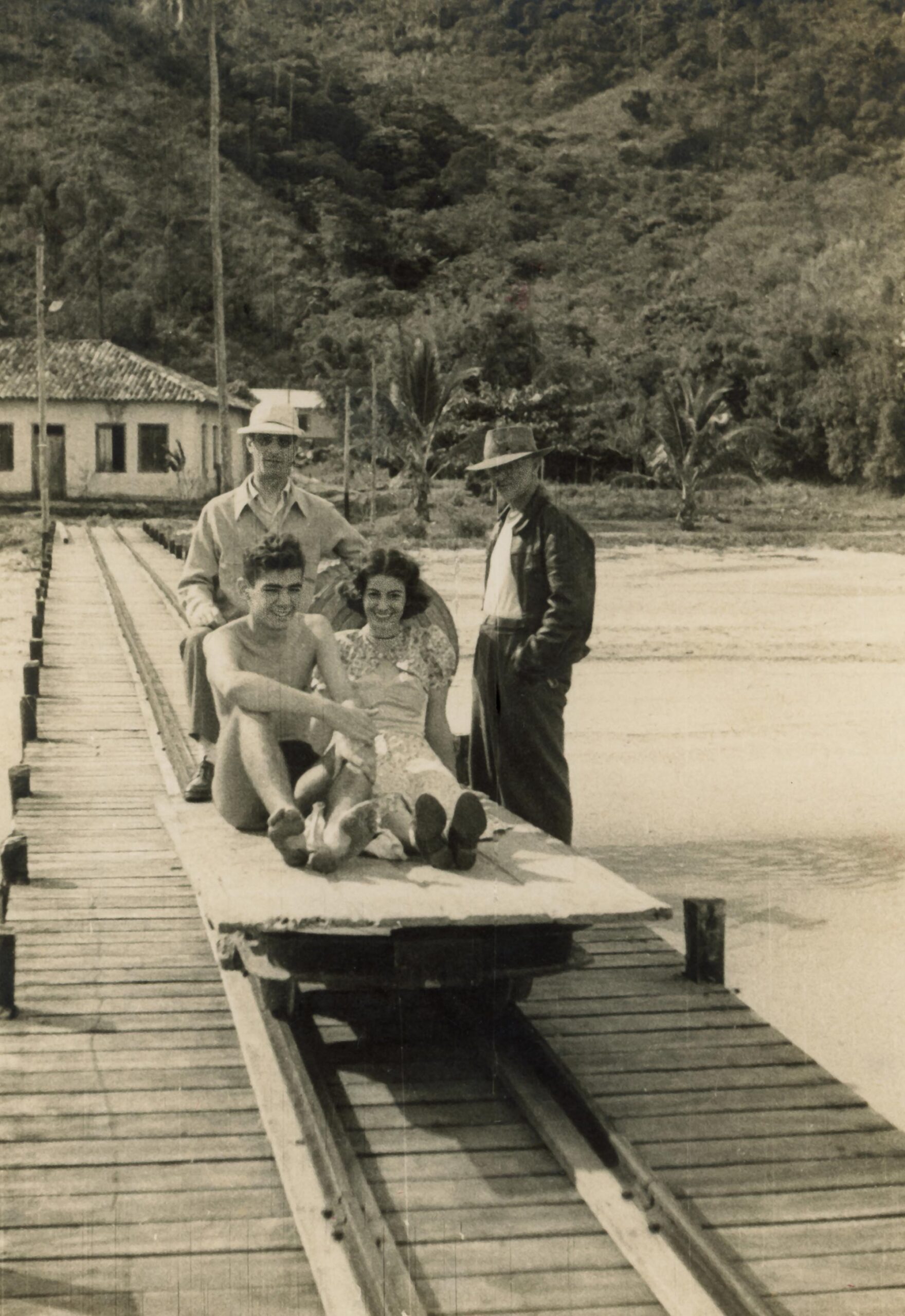 Foto em preto e branco mostra um pequeno carrinho de madeira que est&aacute; em cima de um trilho de trem constru&iacute;do sobre um p&iacute;er em numa faixa de areia. Em cima do carrinho est&aacute; sentada uma jovem mulher sorrindo ao lado de um rapaz sem camisa. Atr&aacute;s deles, de p&eacute;, h&aacute; dois homens de camisa. Ao fundo h&aacute; uma mata e um casar&atilde;o colonial.
