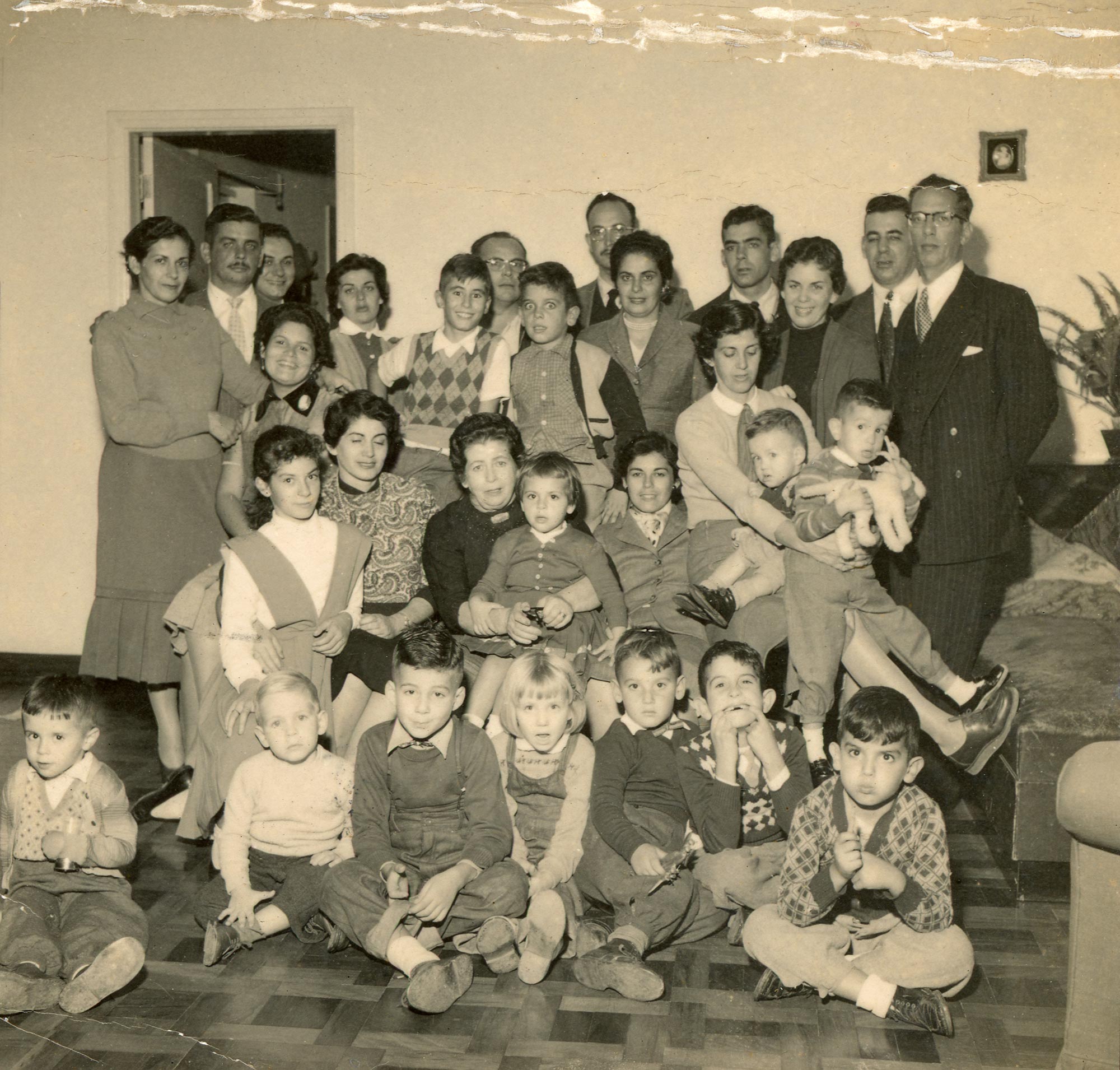 Retrato em preto e branco mostra uma fam&iacute;lia composta por crian&ccedil;as, adultos e idosos posando em um sal&atilde;o.