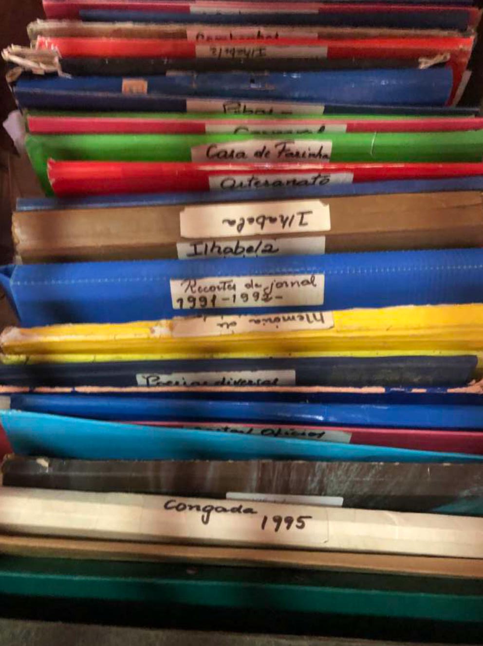 Foto mostra cerca de 15 pastas coloridas alinhadas, algumas delas identificadas com etiquetas escritas a m&atilde;o: Ilhabela, Congada 1995, Recortes de jornal 1991 1992, Casa de Farinha, Artesanato.