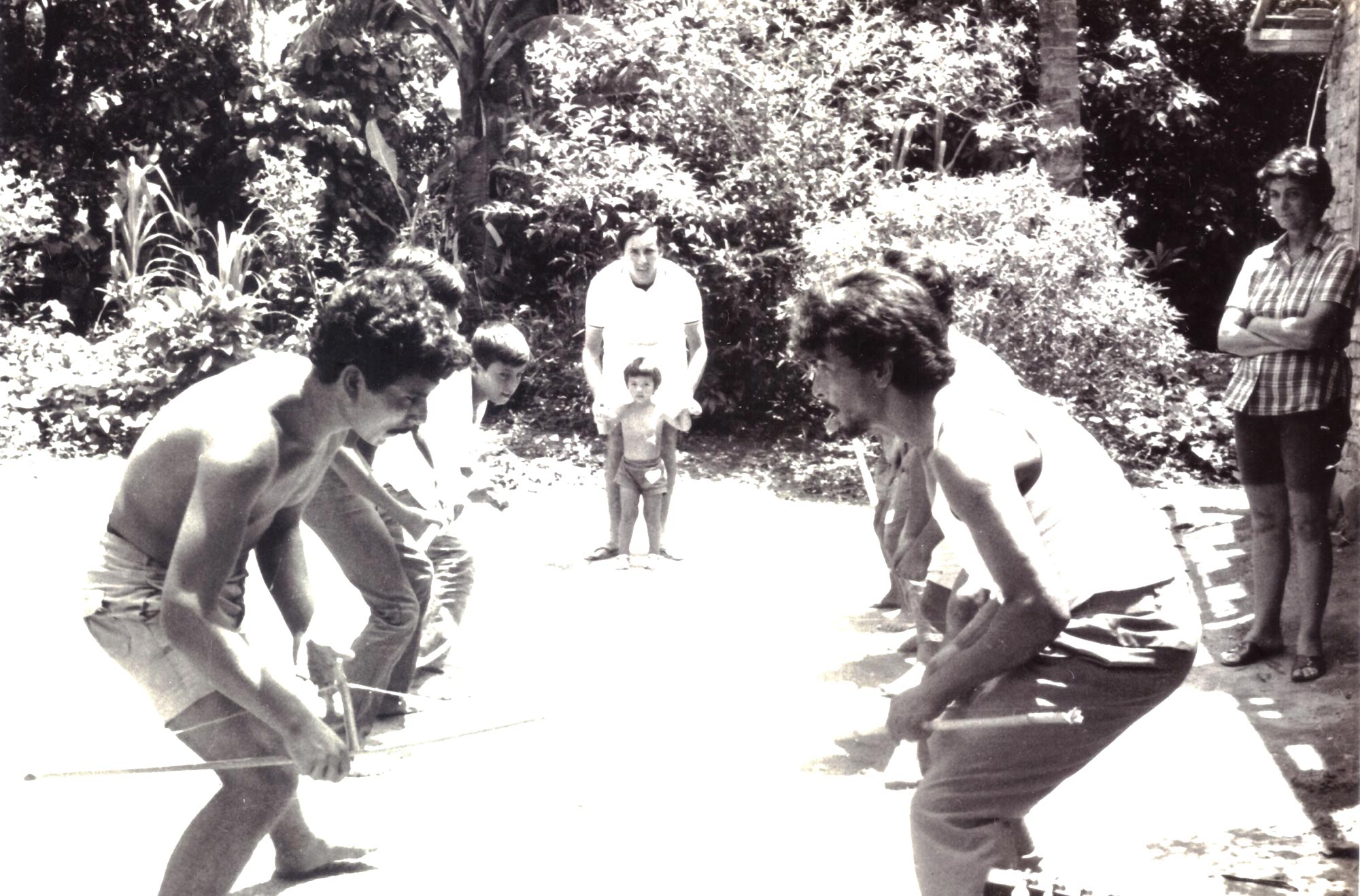 Fotografia em preto e branco mostra o ensaio do caiap&oacute;, com duas fileiras de pessoas alinhadas frente a frente. No primeiro plano h&aacute; um homem sem camisa e de shorts do lado e esquerdo e um homem de camisa e shorts do lado direito. Os dois carregam bodoques, instrumentos semelhantes a um arco e flecha. No canto direito, Ded&eacute; est&aacute; de p&eacute; observando. Ao fundo, entre as duas colunas, um homem de p&eacute; observa acompanhado de uma beb&ecirc;.